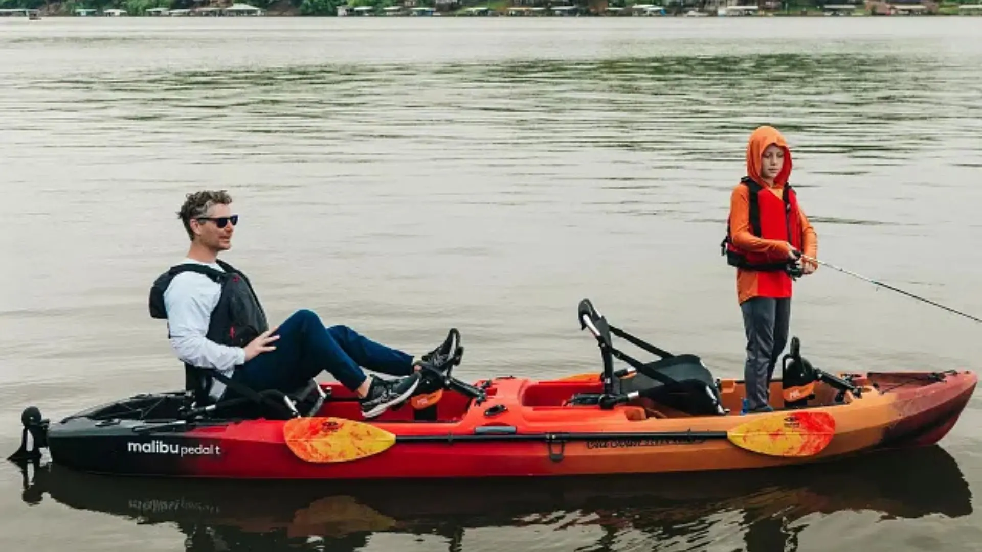 Adult pedaling the Old Town Malibu Pedal T tandem kayak while a child stands and fishes from the front deck.