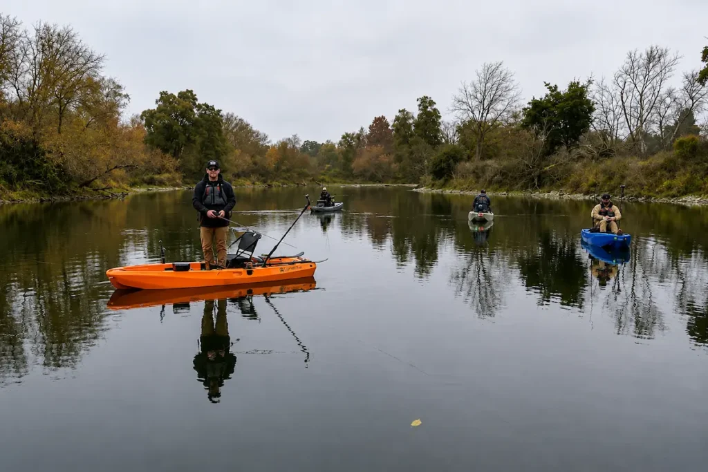 FluxJet Kayak Review: Weed-Ready Electric Jet Power Without an Exposed Prop 1 Four anglers fishing from electric kayaks on a calm tree-lined river, with one angler standing on an orange kayak in the foreground.